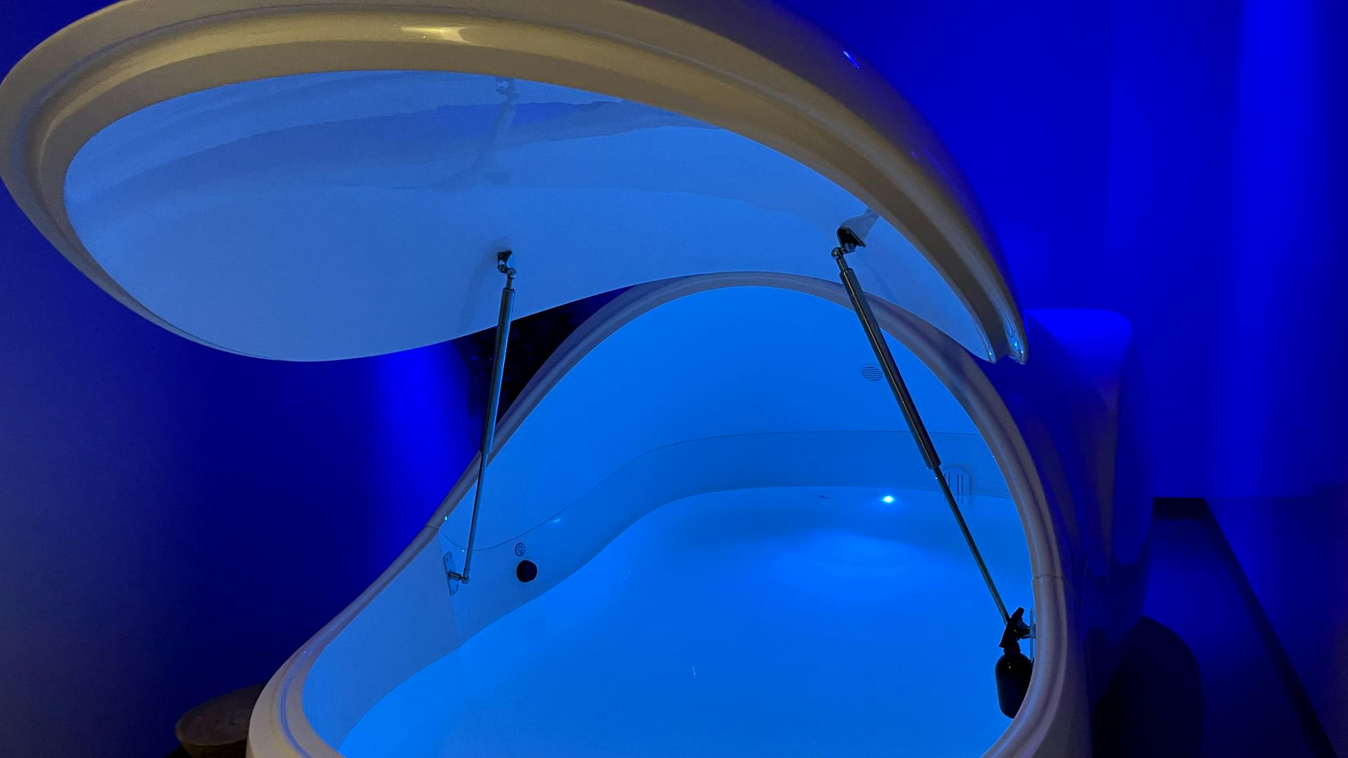 A sensory deprivation tank. The tank is open and it is filled with water. The tank is lit by light blue light.